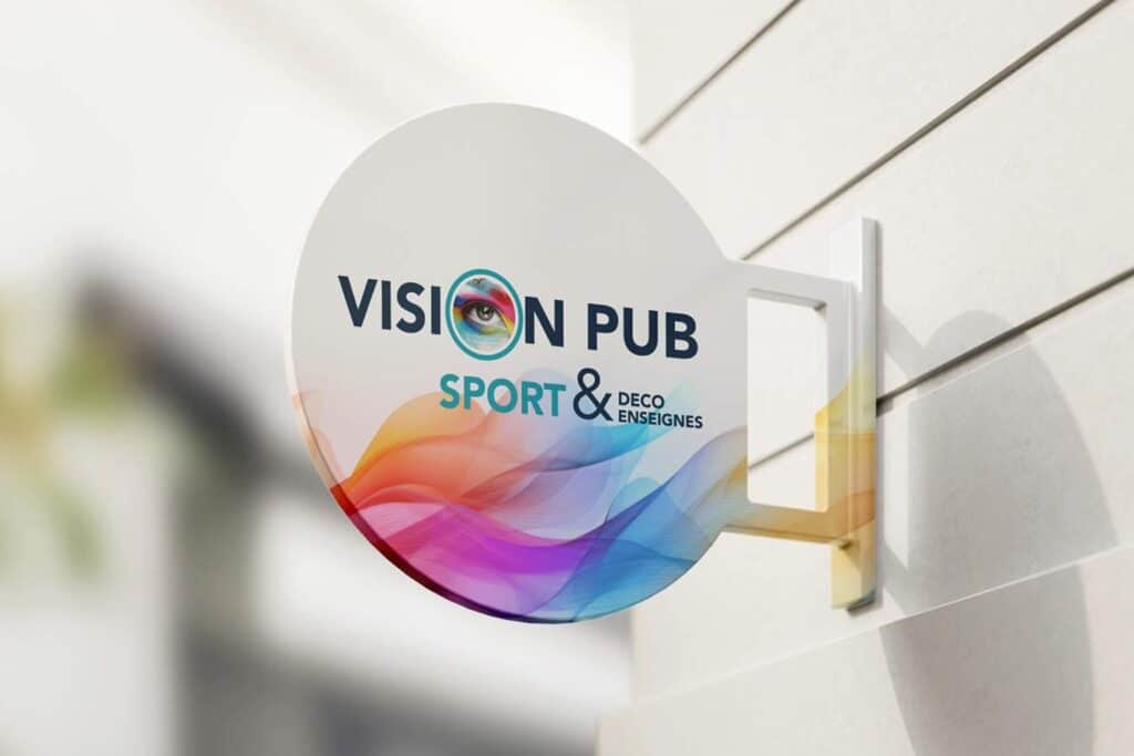 Panneau rond blanc "VISION PUB" avec un œil coloré, "SPORT & DECO ENSEIGNES" en bas. Accroché à un mur extérieur clair.