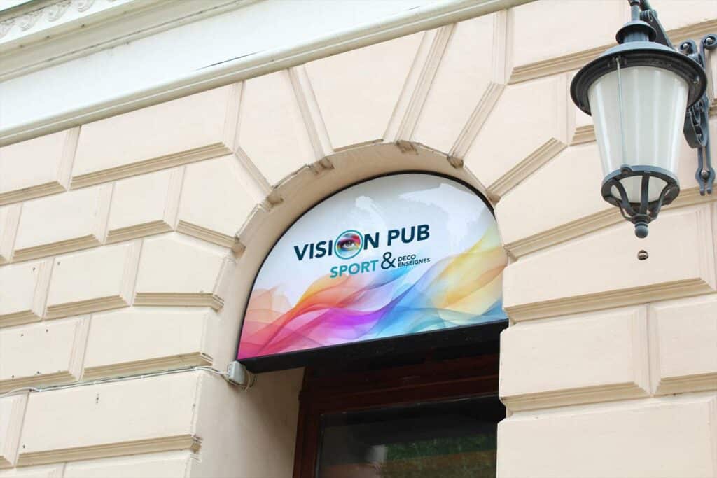 Enseigne semi-circulaire lumineuse "VISION PUB SPORT & DECO ENSEIGNES" sur façade en pierre avec motifs, près d'un lampadaire. Design coloré.