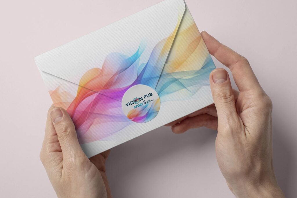 Deux mains tiennent une enveloppe texturée aux motifs ondulés et colorés. Un autocollant rond "VISION PUB SPORT & DECO PASSIONS" est apposé au centre.