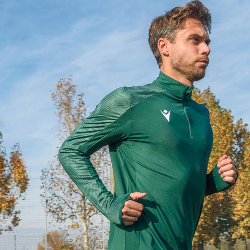 Homme concentré courant, portant un maillot de running Macron vert foncé à manches longues avec zip. Fond d'arbres automnaux et ciel bleu.