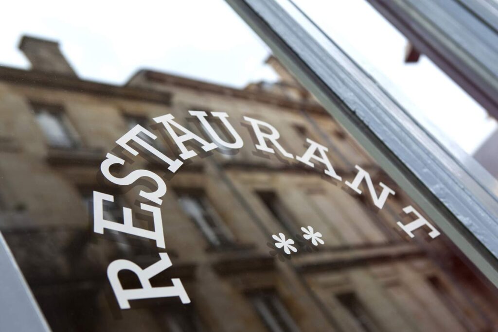 Enseigne "RESTAURANT" blanche courbée sur une vitrine sombre avec deux étoiles. Reflet d'un bâtiment ancien en arrière-plan flou.