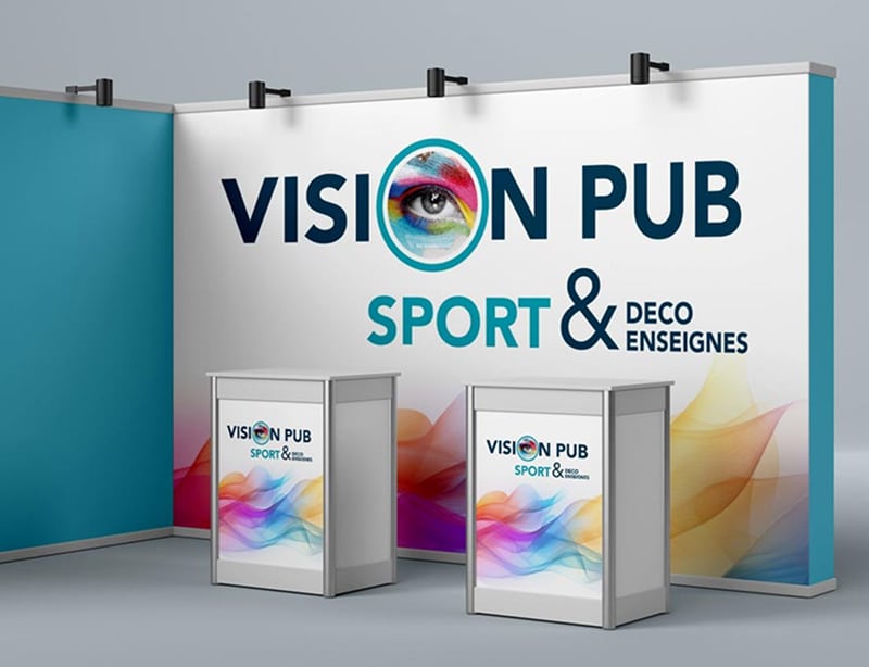 Stand d'exposition Vision Pub Sport & Déco Enseignes: grand panneau avec logo œil coloré, texte, et deux comptoirs d'accueil assortis.