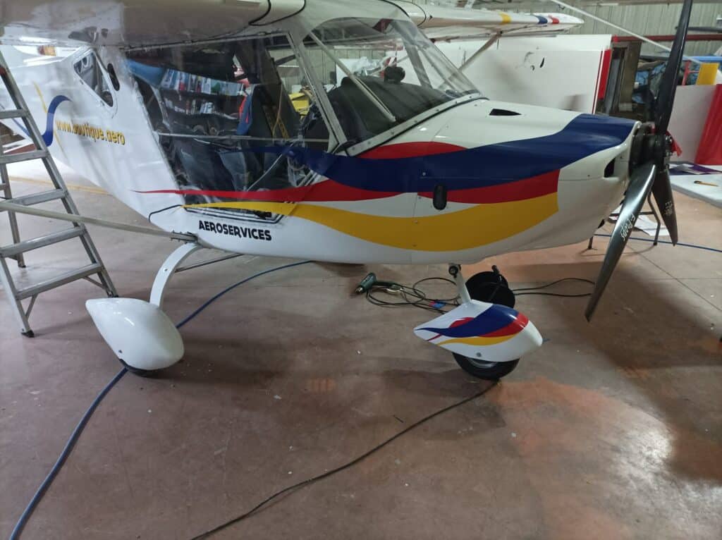 Avion blanc à rayures bleu, rouge, jaune, texte "AEROSERVICES", hélice "E-Props", et "www.boutique.aero". En maintenance hangar.