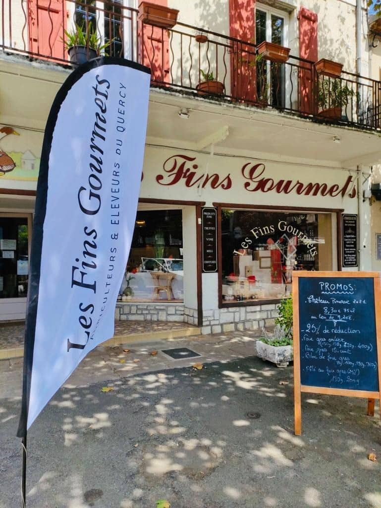 Façade de "Les Fins Gourmets" au Quercy avec drapeau, pancarte "PROMOS" (foie gras, château Pinerie) et détails architecturaux.