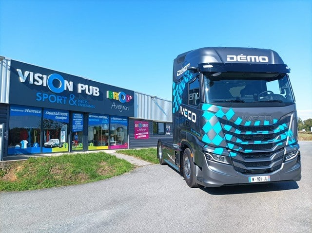 Camion Iveco gris avec motif à carreaux bleus 'DÉMO' devant un bâtiment 'VISION PUB BROD' Aveyron', spécialiste marquage véhicule, enseignes.