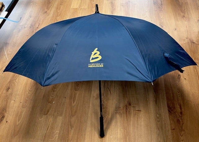 Parapluie ouvert bleu marine avec logo jaune B et texte "La Boutique du Menuisier" posé sur un plancher en bois.