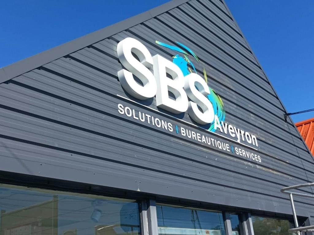 Enseigne SBS Solutions Bureautique Services Aveyron sur façade grise. Logo blanc 3D avec motifs bleu et vert.