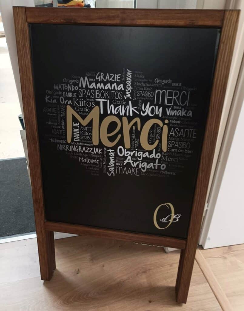Tableau noir "Merci" : Un message de gratitude multilingue Tableau noir en bois foncé avec un nuage de mots en forme de cœur. Le mot "Merci" doré est central, entouré de "Thank you" en plusieurs langues. Logo OB en bas.