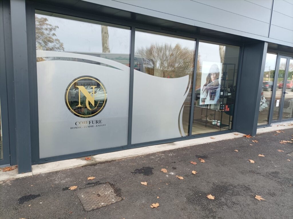 Coiffure Homme Femme Enfant : Logo Doré sur Vitrine Façade d'un salon de coiffure pour homme, femme, enfant avec logo doré 'N' et vitres dépolies, devant un parking d'automne.